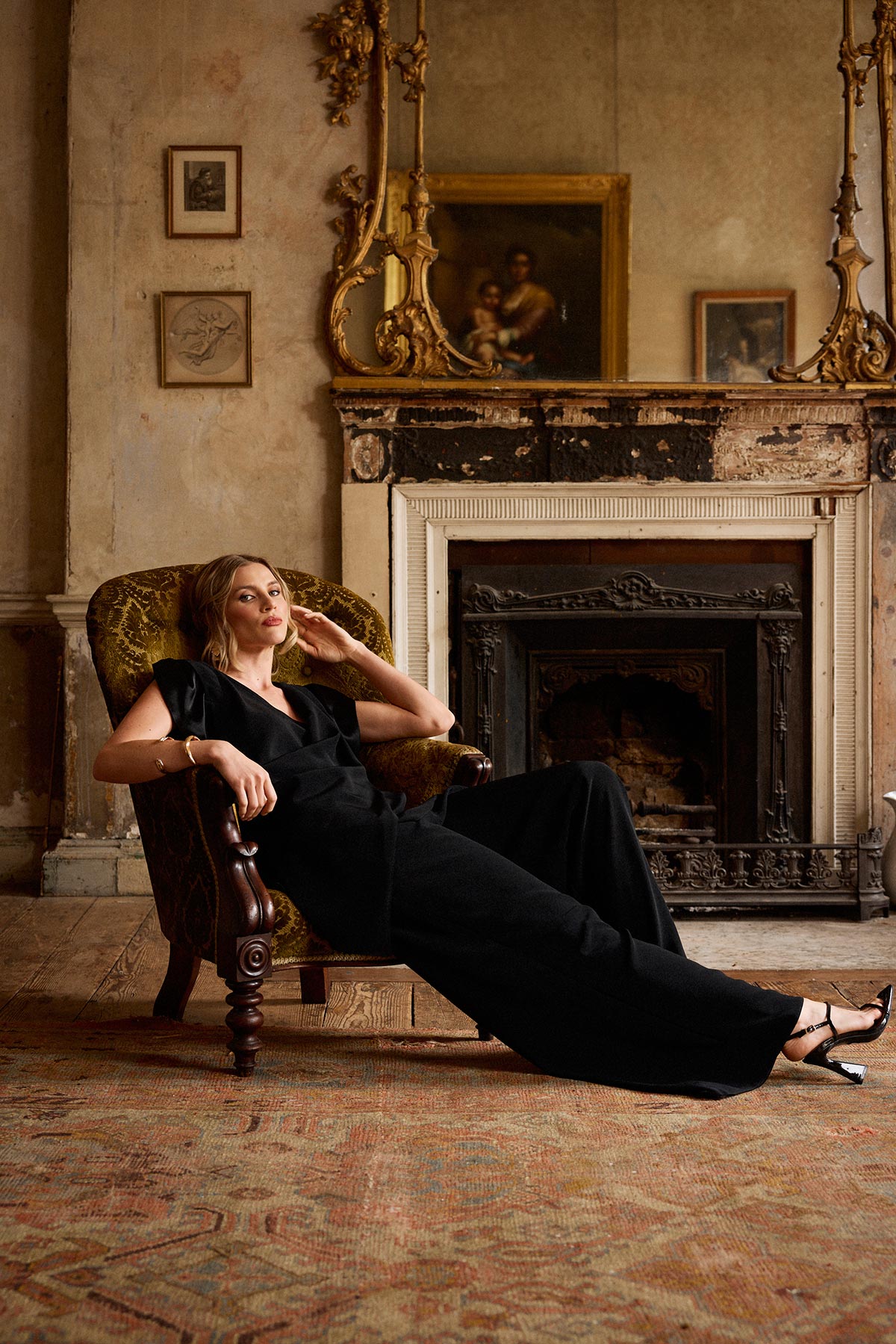 Woman in a black outfit sitting in an ornate chair in a room with a fireplace and decorative mirror.
