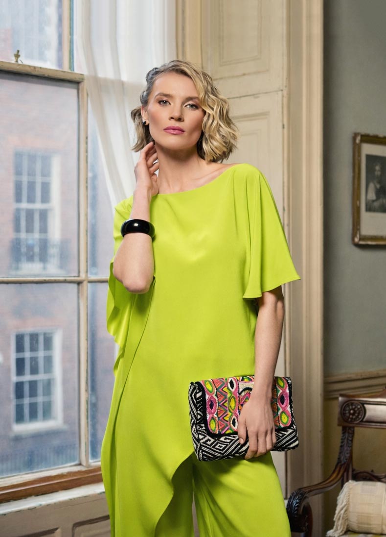 Woman in a bright green outfit holding a colorful clutch in a room with a window.