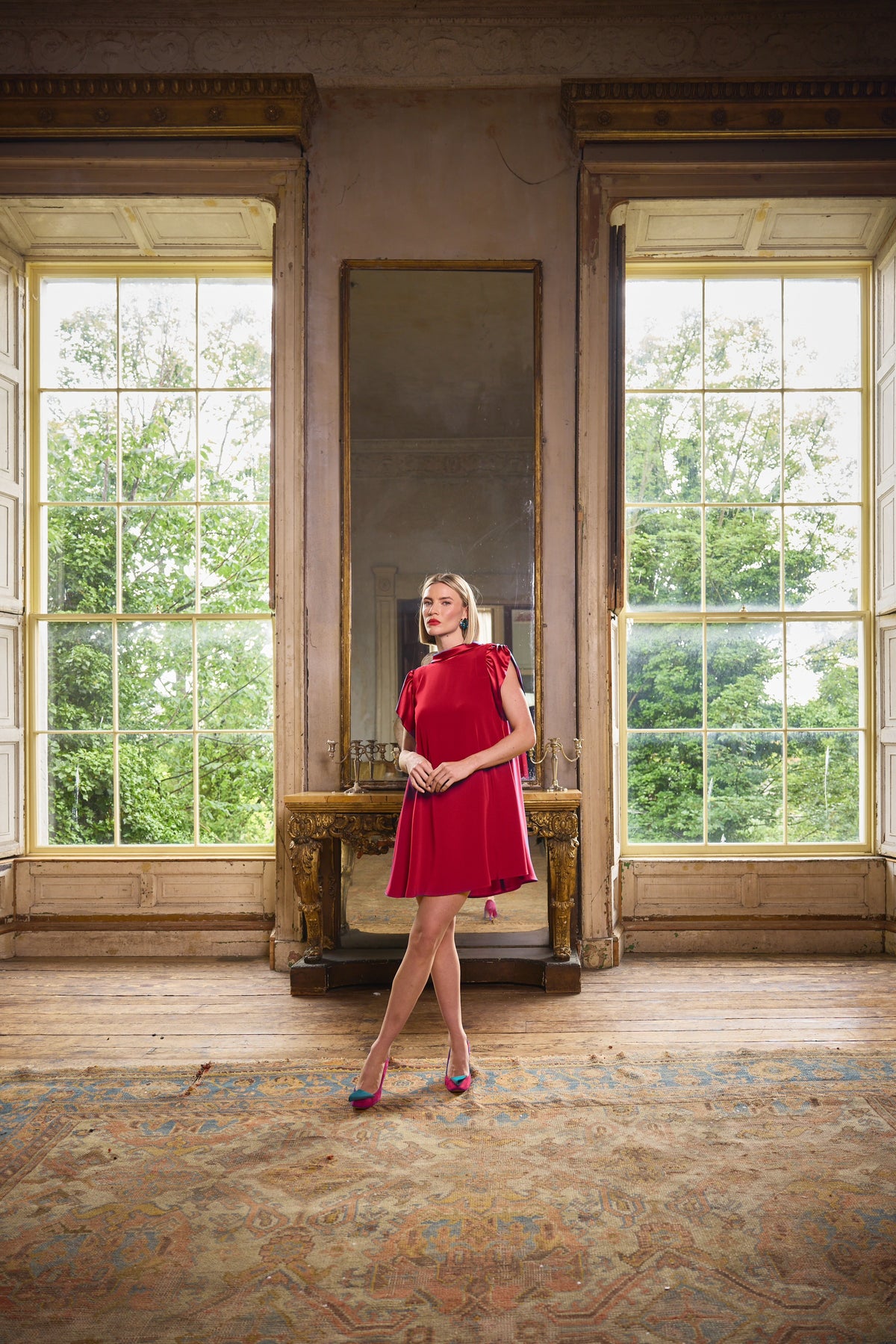 A woman wearing a bright red sleeveless dress and colorful heels stands in a large, sunlit room with tall windows, a mirror, and a patterned rug.