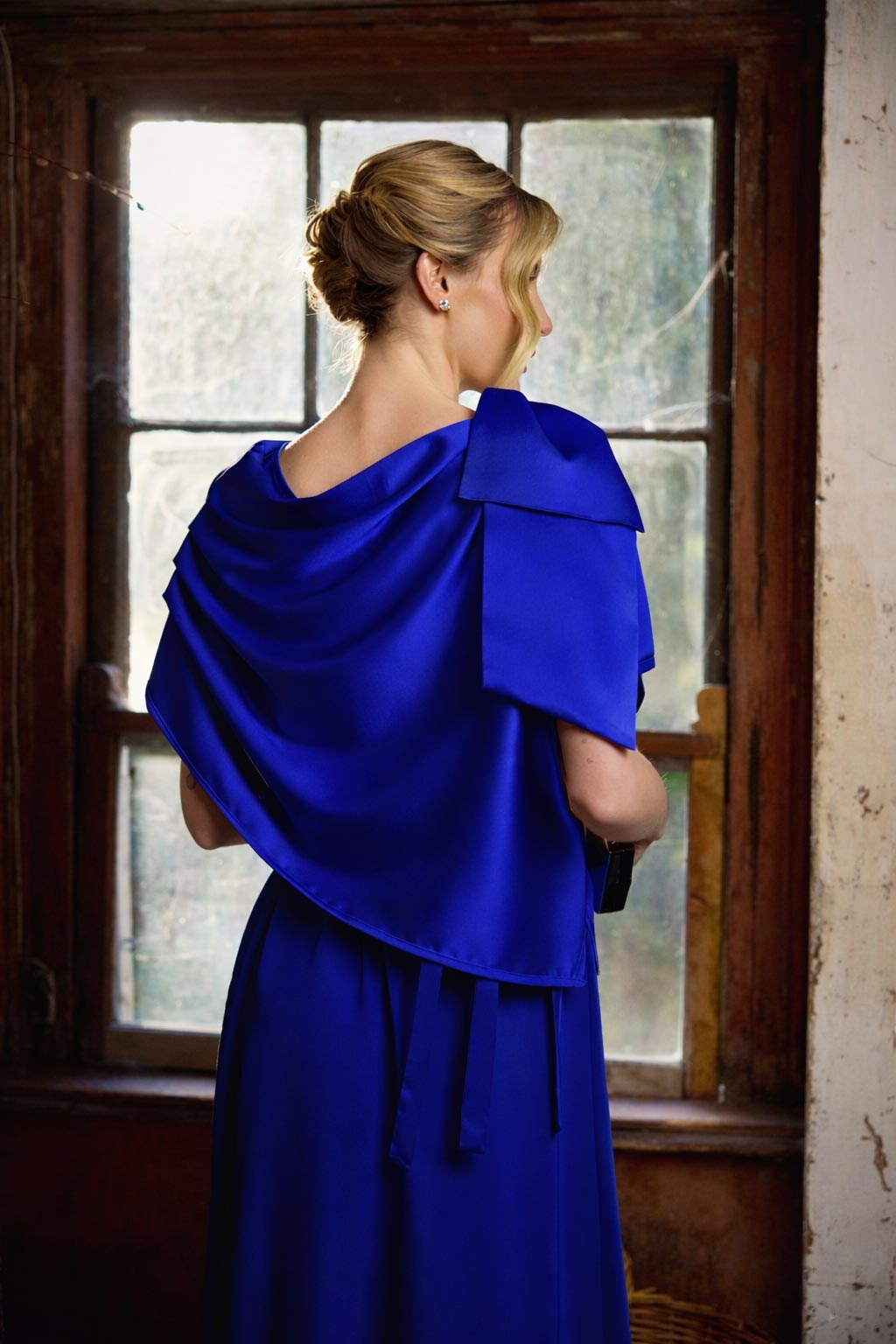 Woman wearing a blue dress with a draped effect in front of a window.
