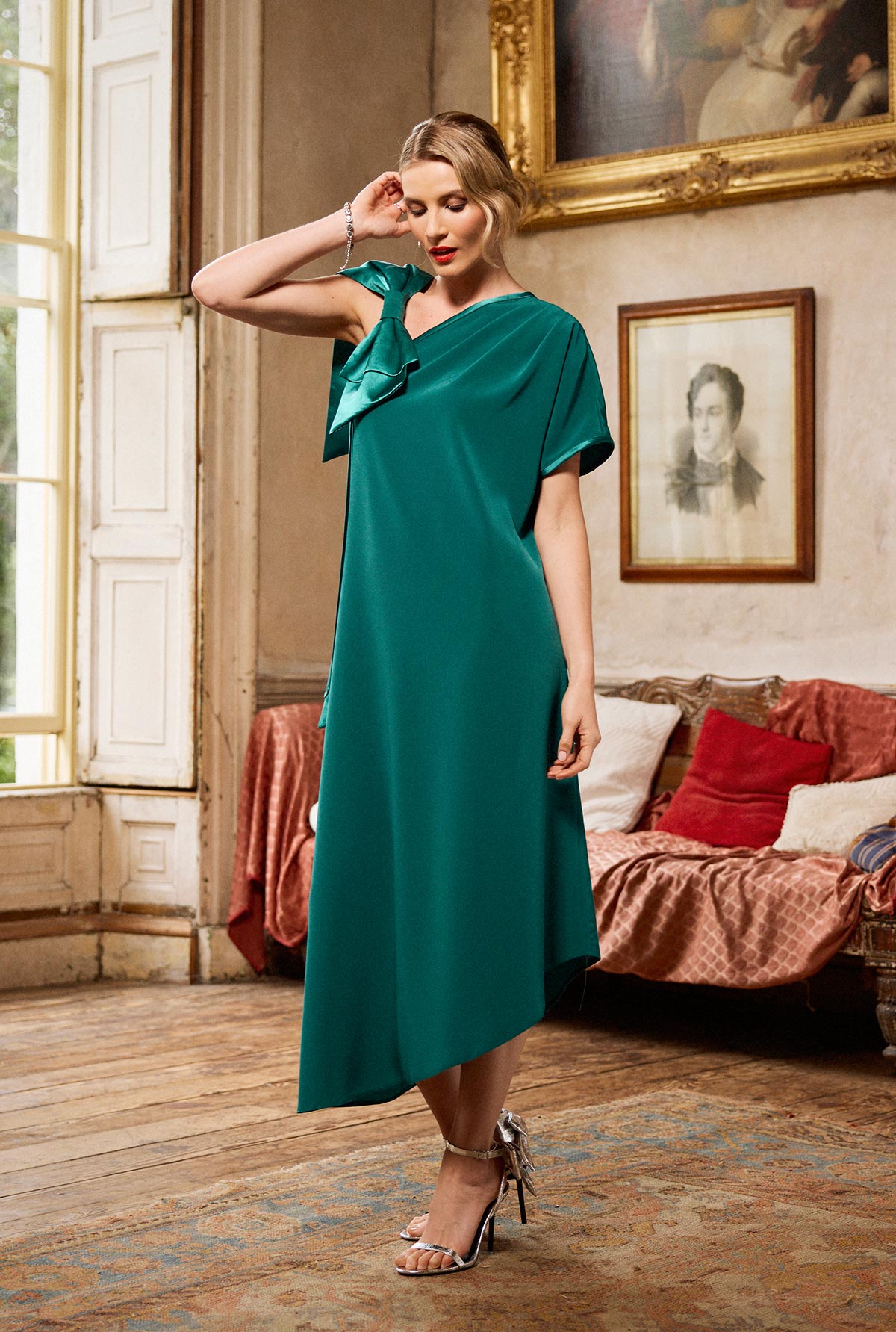 Woman in a green dress standing in an elegant room with a couch and framed pictures.