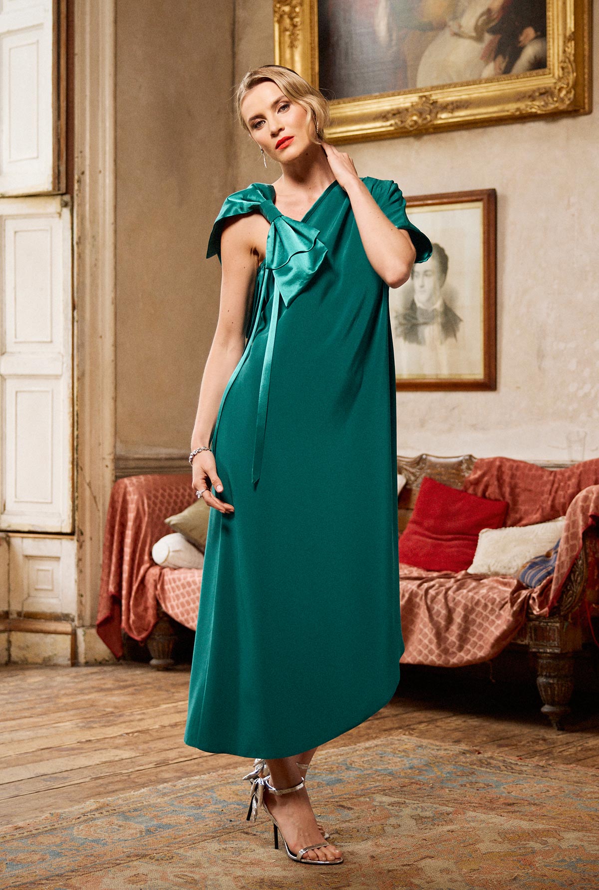 Woman in a teal dress standing in an elegant room with a painting and sofa in the background.