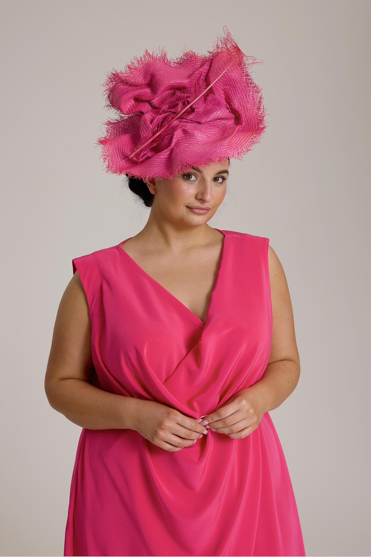 Person in a bright pink dress and an elaborate pink hat with a feathery design, standing against a plain background.