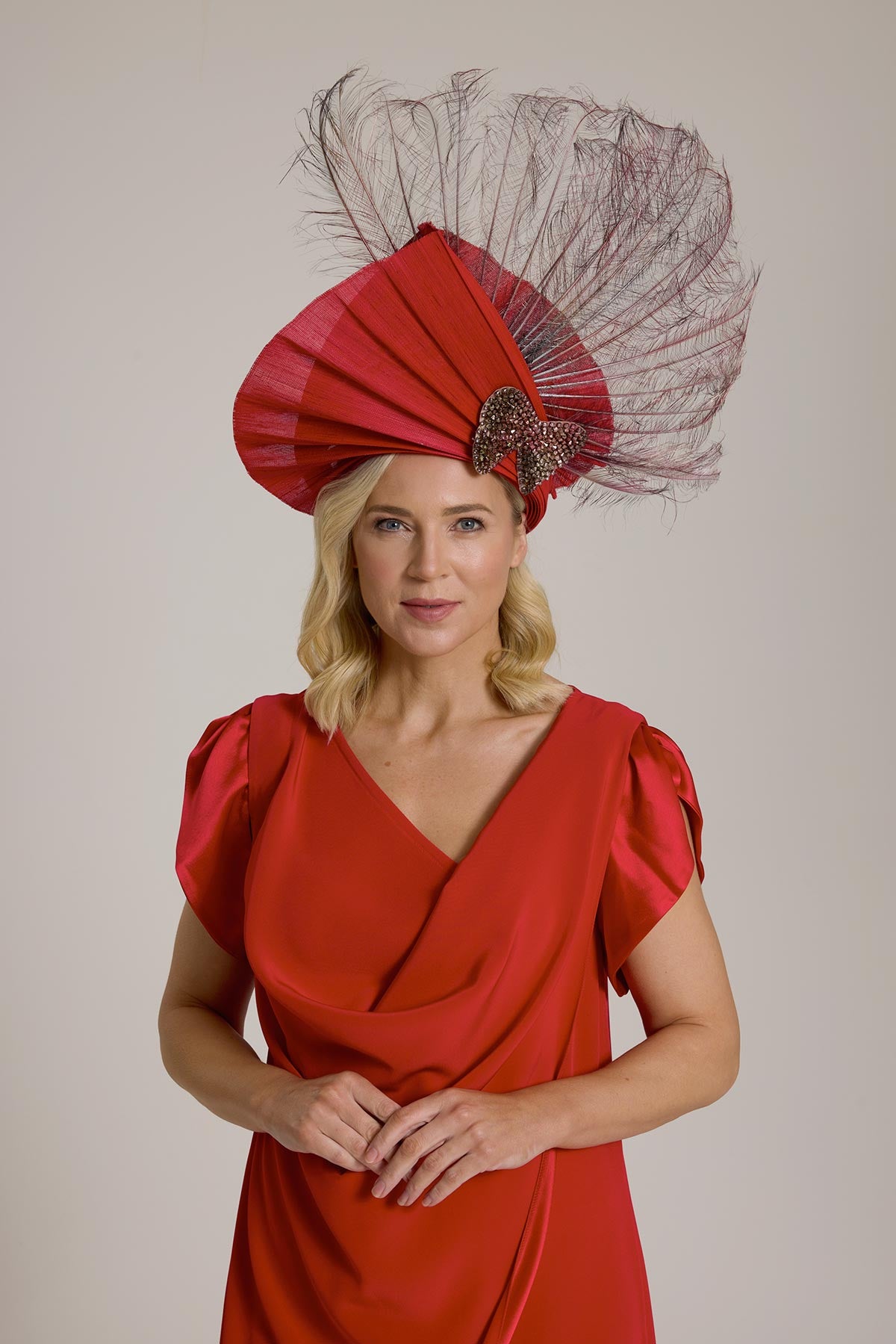 A woman in a long red dress and a large, feathered hat, standing against a plain background.