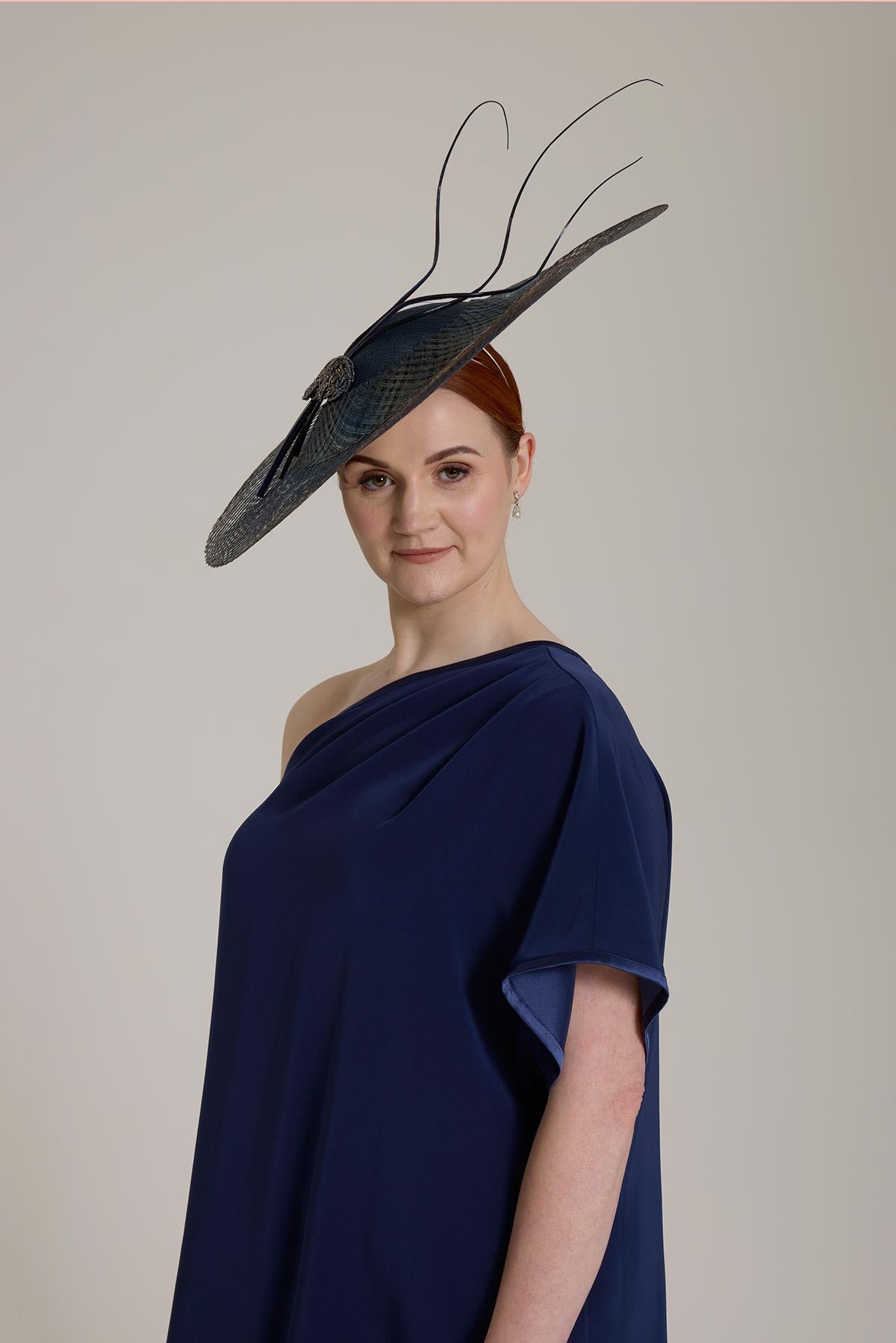 Woman in navy dress and dramatic hat with feathers against a neutral background.