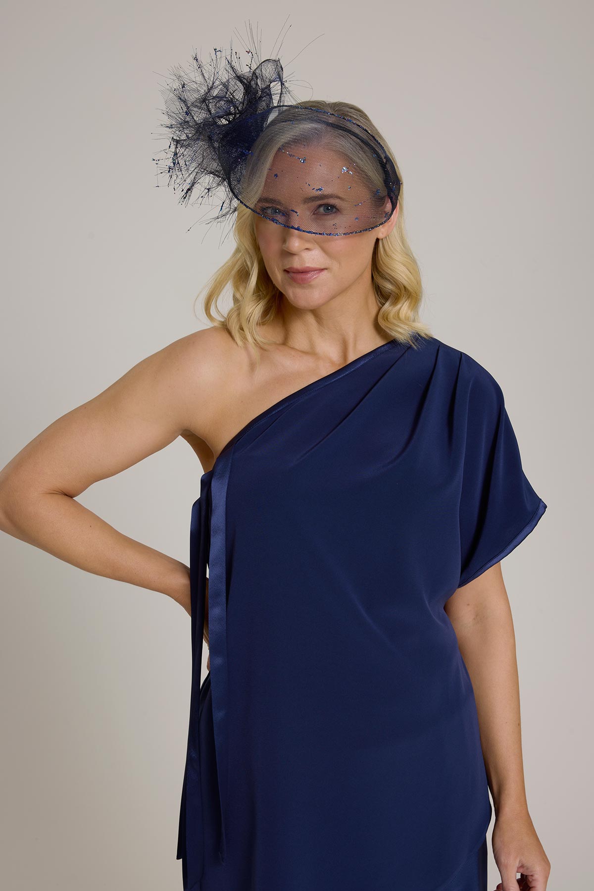 A model in a navy one-shoulder dress wearing a matching netted headpiece.