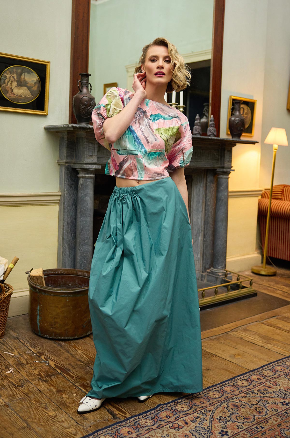 Woman in a colorful top and teal skirt standing in an elegant room with a fireplace and decorative items.