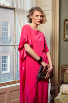 Woman in a bright pink dress standing in a room with a window and chair.