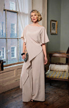 Woman in a beige dress standing in a room with large windows and a chair.