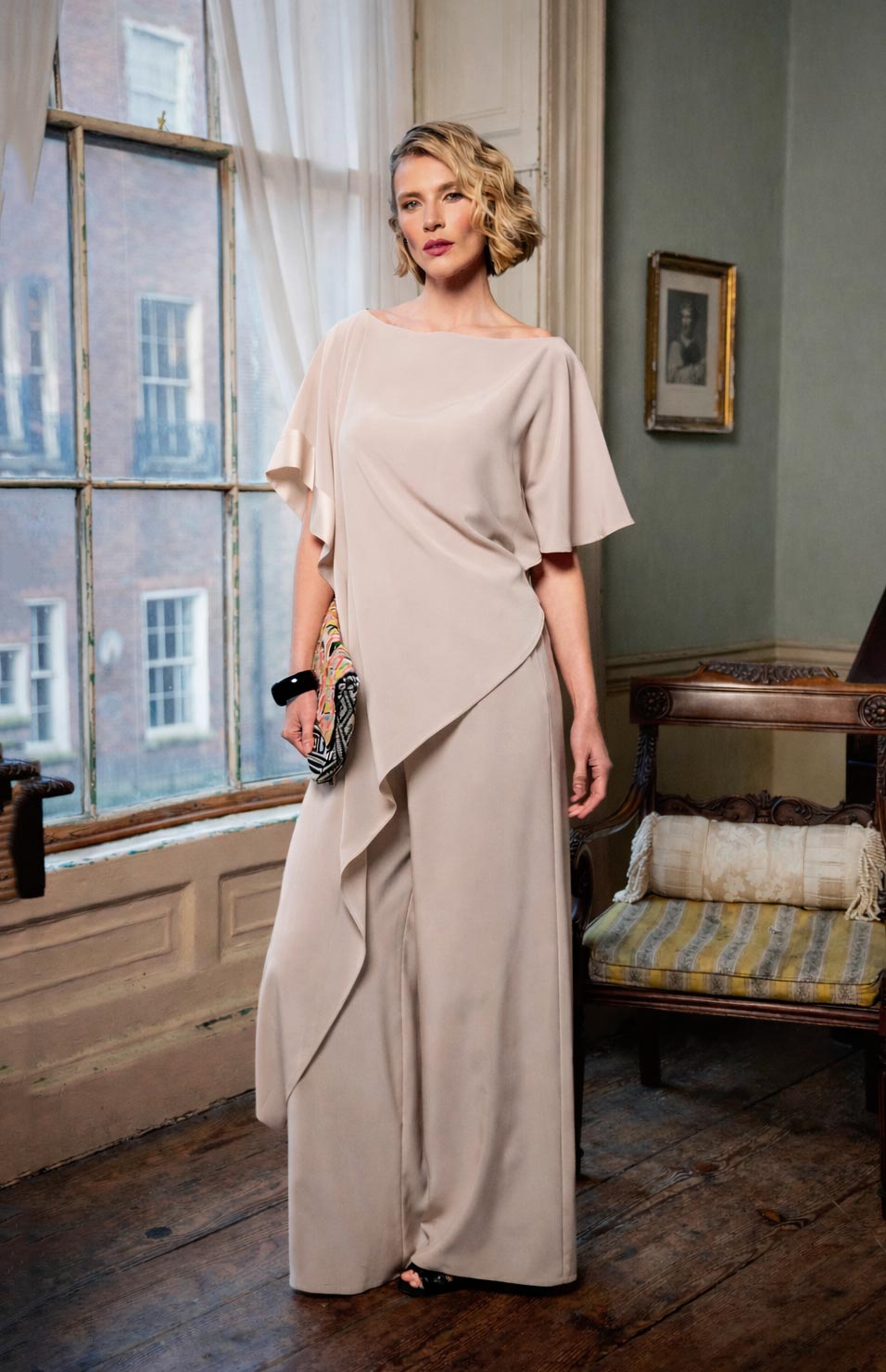 Woman in a beige dress standing in a room with large windows and a chair.