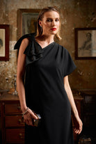Woman in a black dress with a shoulder  bow standing in an ornate room with framed pictures on the wall.