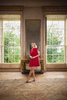 A woman wearing a bright red sleeveless dress and colorful heels stands in a large, sunlit room with tall windows, a mirror, and a patterned rug.