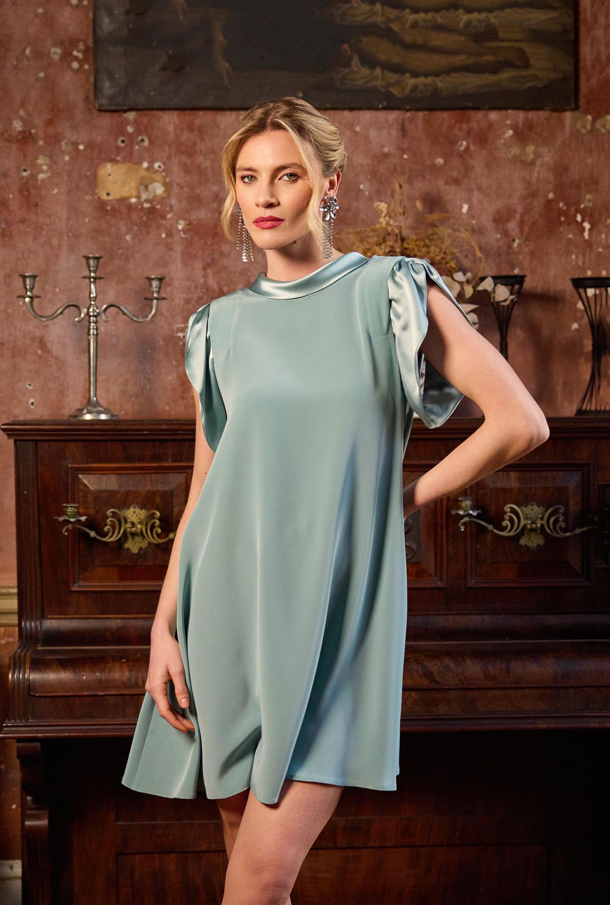 Woman in a light blue dress standing in an elegant room with wooden furniture and decorative elements.