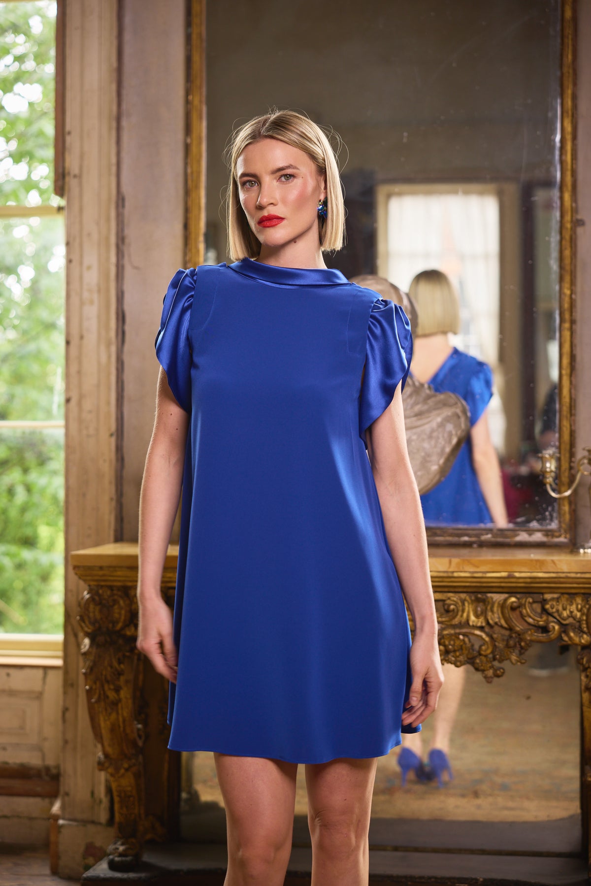 A woman wearing a bright royal blue short-sleeve dress with puffed cap sleeves, standing in a vintage room with a large ornate mirror behind her.