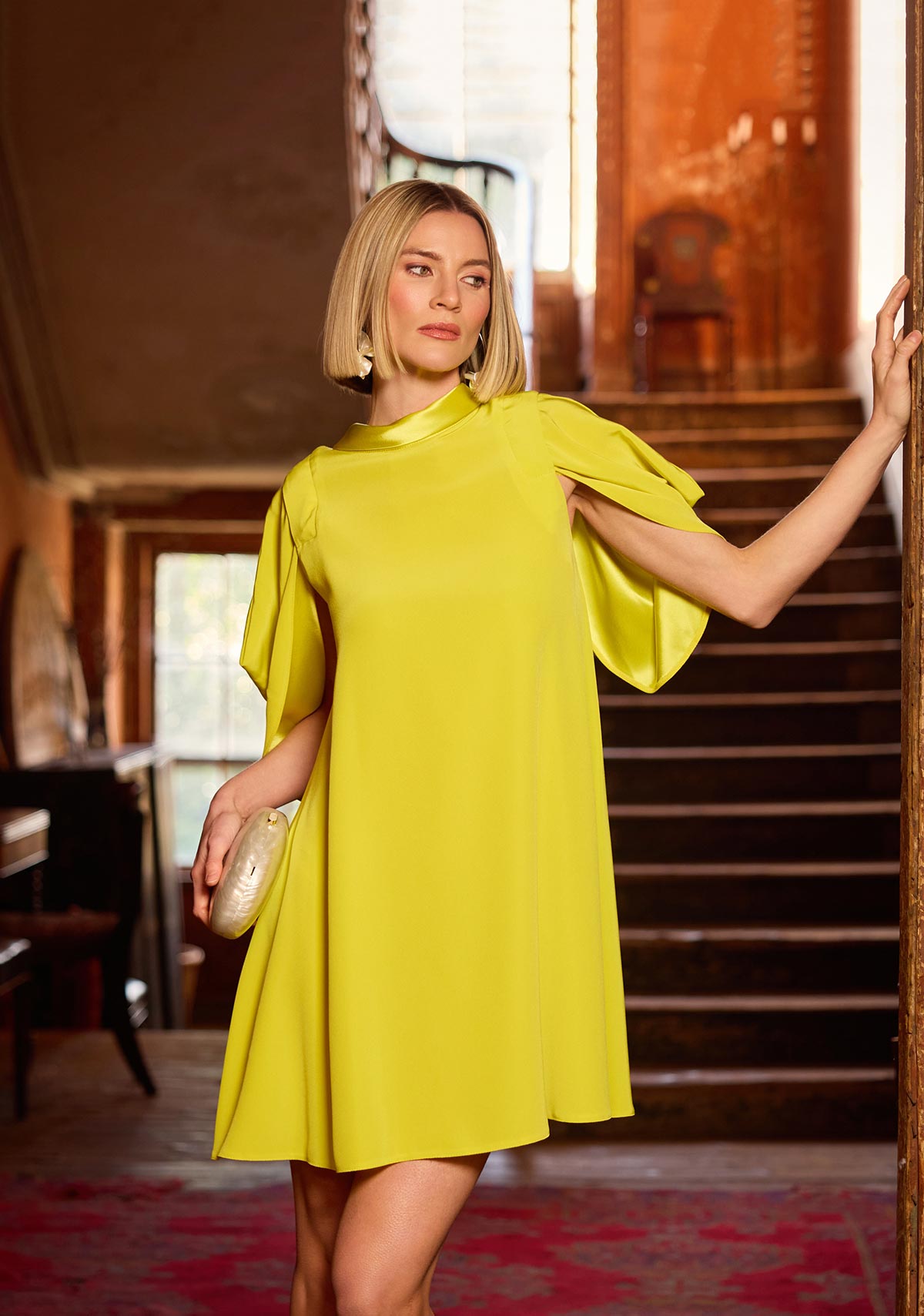 Woman in a yellow dress standing in an indoor setting with wooden stairs and decor.