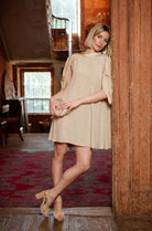 Woman in a beige dress standing in an indoor setting with wooden walls and a rug.