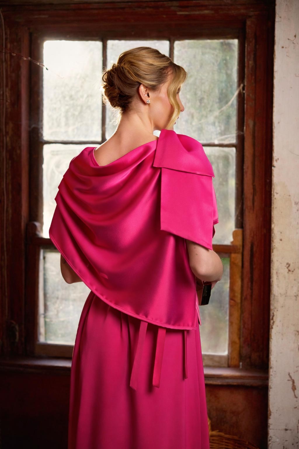 Woman wearing a bright pink dress with a draped effect, standing in front of a window.