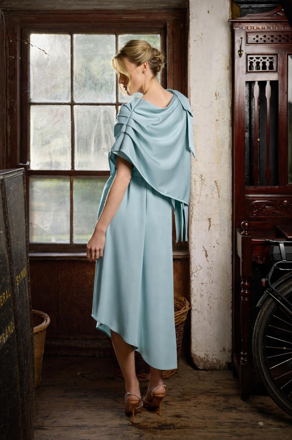 Woman in a light blue dress with a cape standing in a room with wooden furniture and a window.