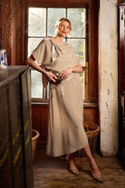 Woman in a beige dress standing in a rustic room with wooden floors and a large window.