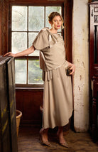 Woman in a beige dress standing in a room with a window and wooden furniture.