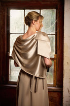 Woman wearing a beige satin shawl over a dress, standing in front of a window.