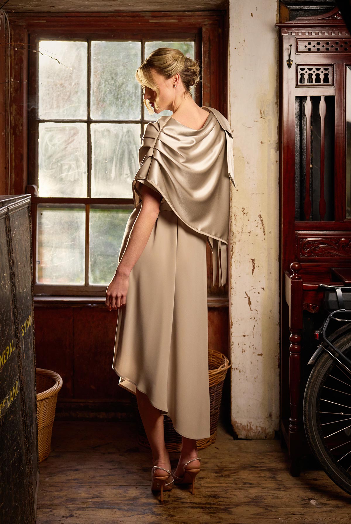 Woman in a beige dress and cape standing in a room with wooden furniture and a bicycle.