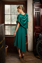 Woman in a teal dress standing in an indoor setting with a window and vintage furniture.