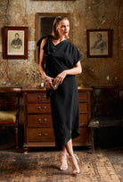 Woman in a black dress standing in an ornate room with wooden furniture and portraits.