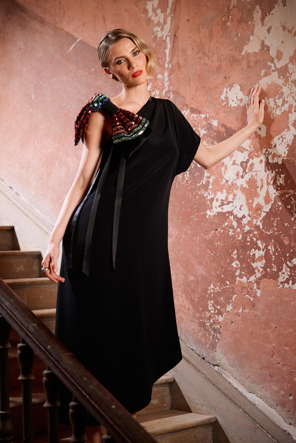 Woman in a black dress with colorful accents standing on a staircase against a textured wall.