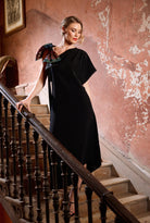 Woman in a black dress holding a colorful shoulder bow on a staircase with a pink wall.