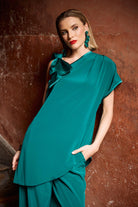 Woman wearing a teal top and trousers against a textured brown wall