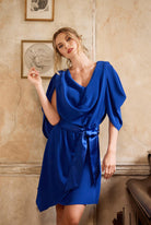 Woman wearing a blue dress standing against a beige wall with framed pictures.