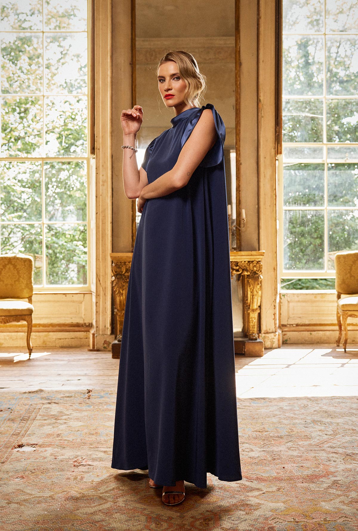 Woman in a long navy dress standing in an elegant room with large windows.