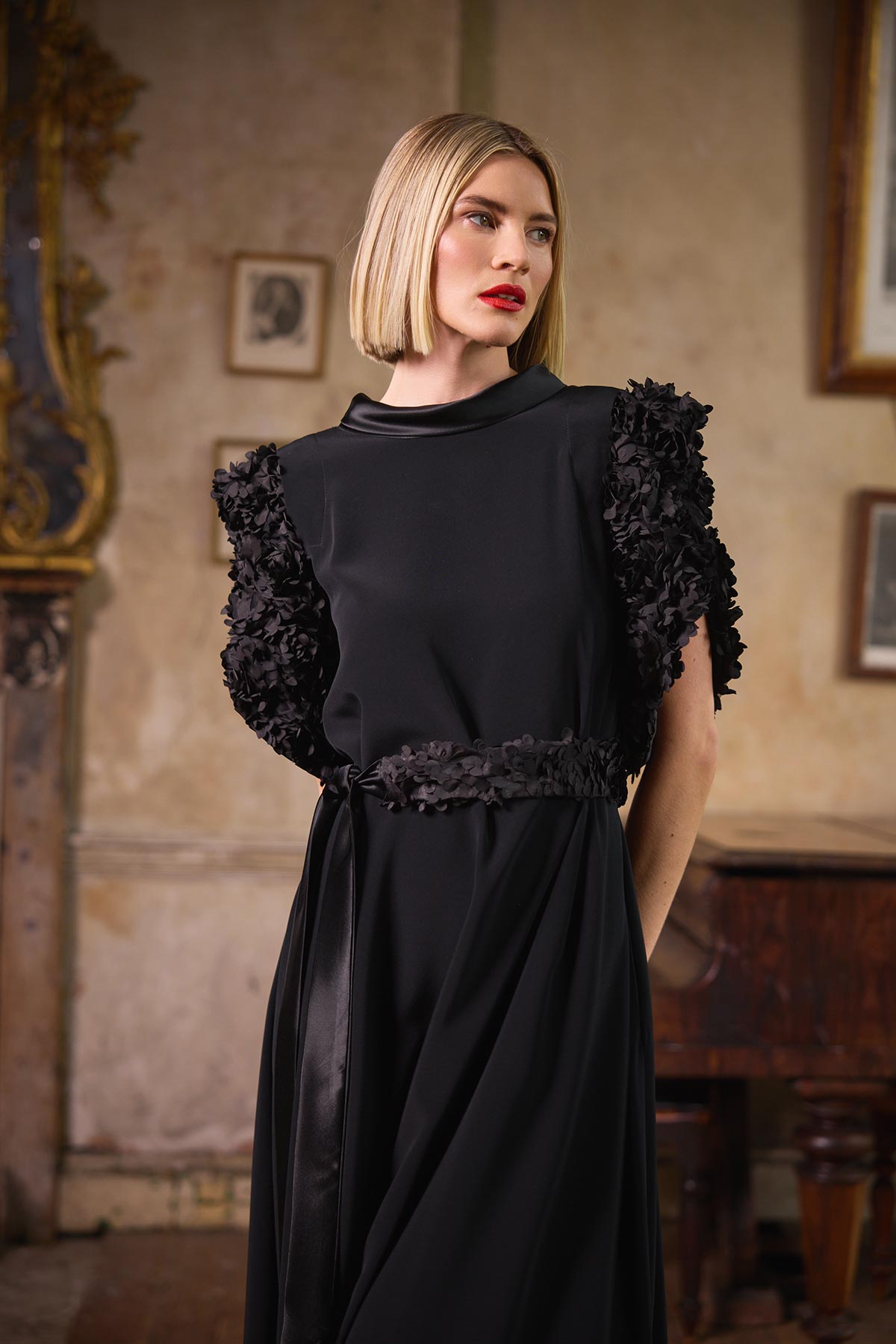 A woman with a short blonde bob wearing a black midi dress with dramatic ruffled cap sleeves and a matching ruffled waistband, standing in an elegant, vintage-style room with warm beige walls and framed pictures.