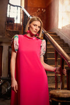 Woman in a bright pink dress standing in an elegant interior setting with stairs and railings.