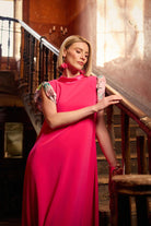 Woman in a bright pink dress standing in an elegant interior setting with a staircase.