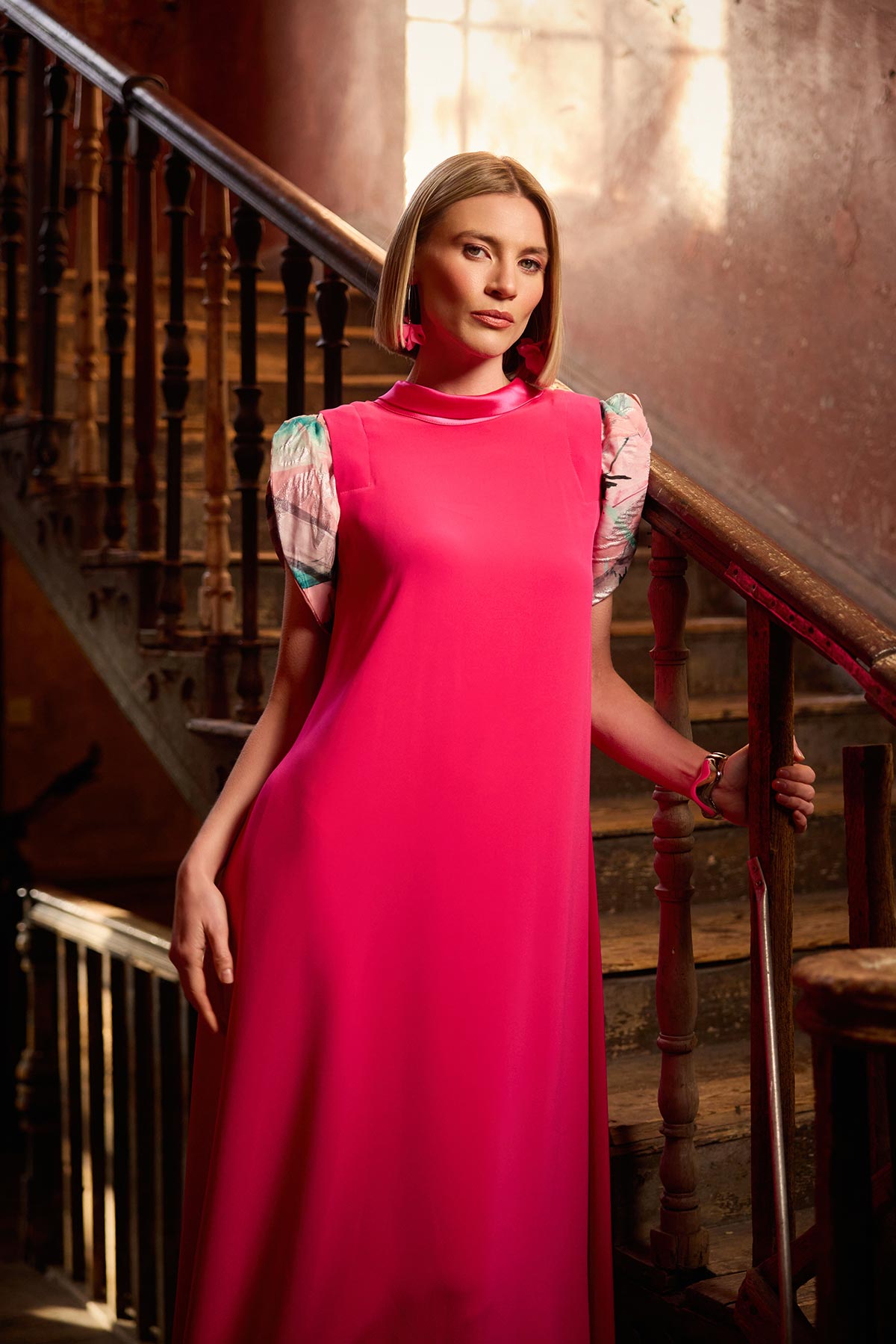 Woman in a pink dress standing in an elegant interior setting with wooden staircase and warm lighting.