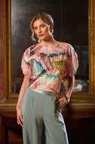 Woman wearing a colorful blouse and light gray pants in front of an ornate mirror.