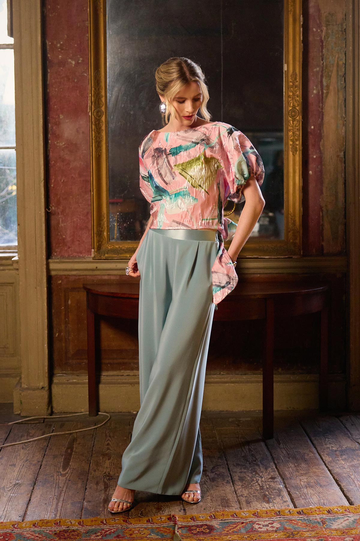 Woman in a colorful top and light green skirt standing in an ornate room with large artwork on the wall.