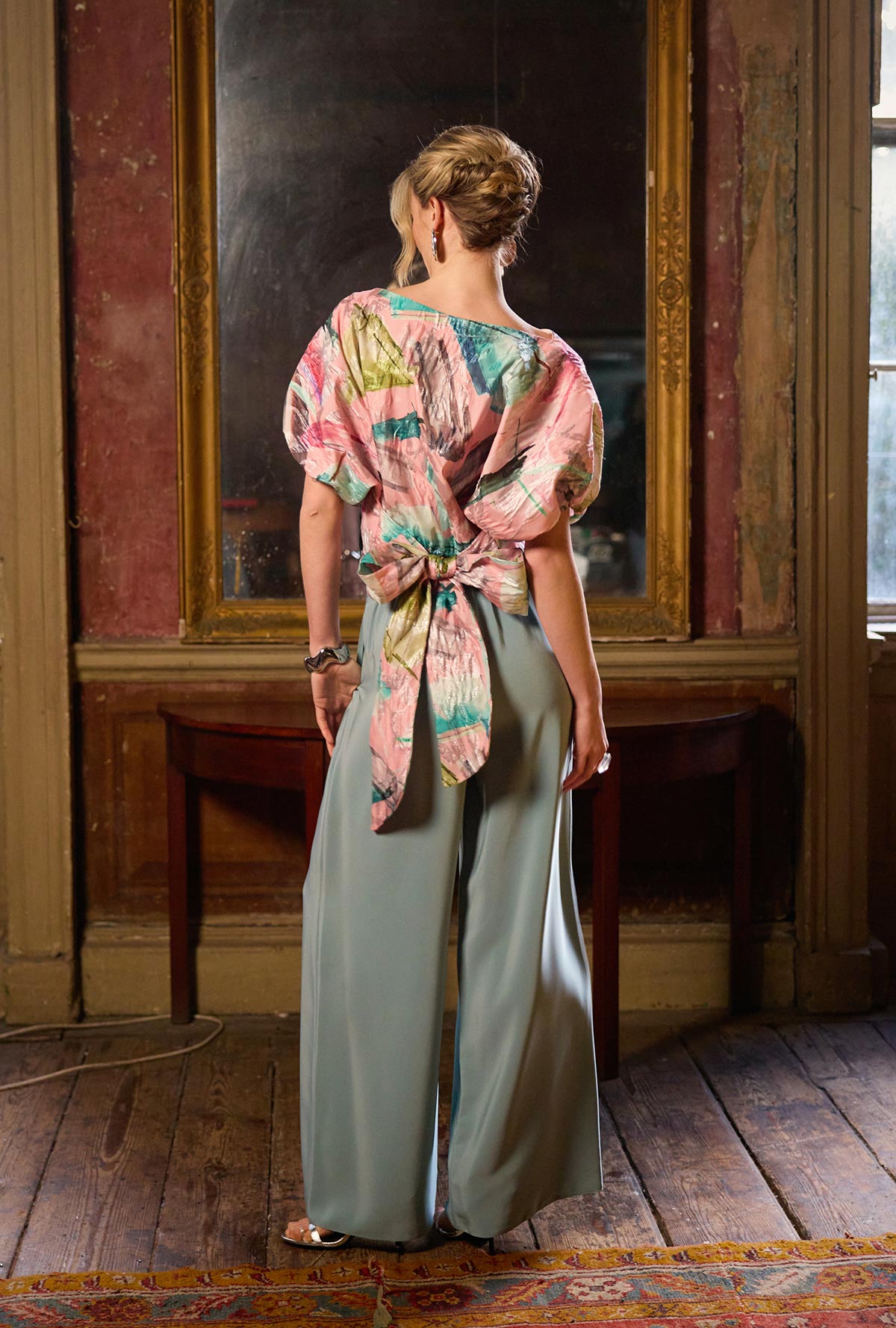 Woman in a colorful top and light green pants standing in an ornate room with wooden floors and a large mirror.