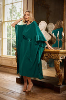 Woman in a green dress standing in an elegant room with large windows and classical decor.