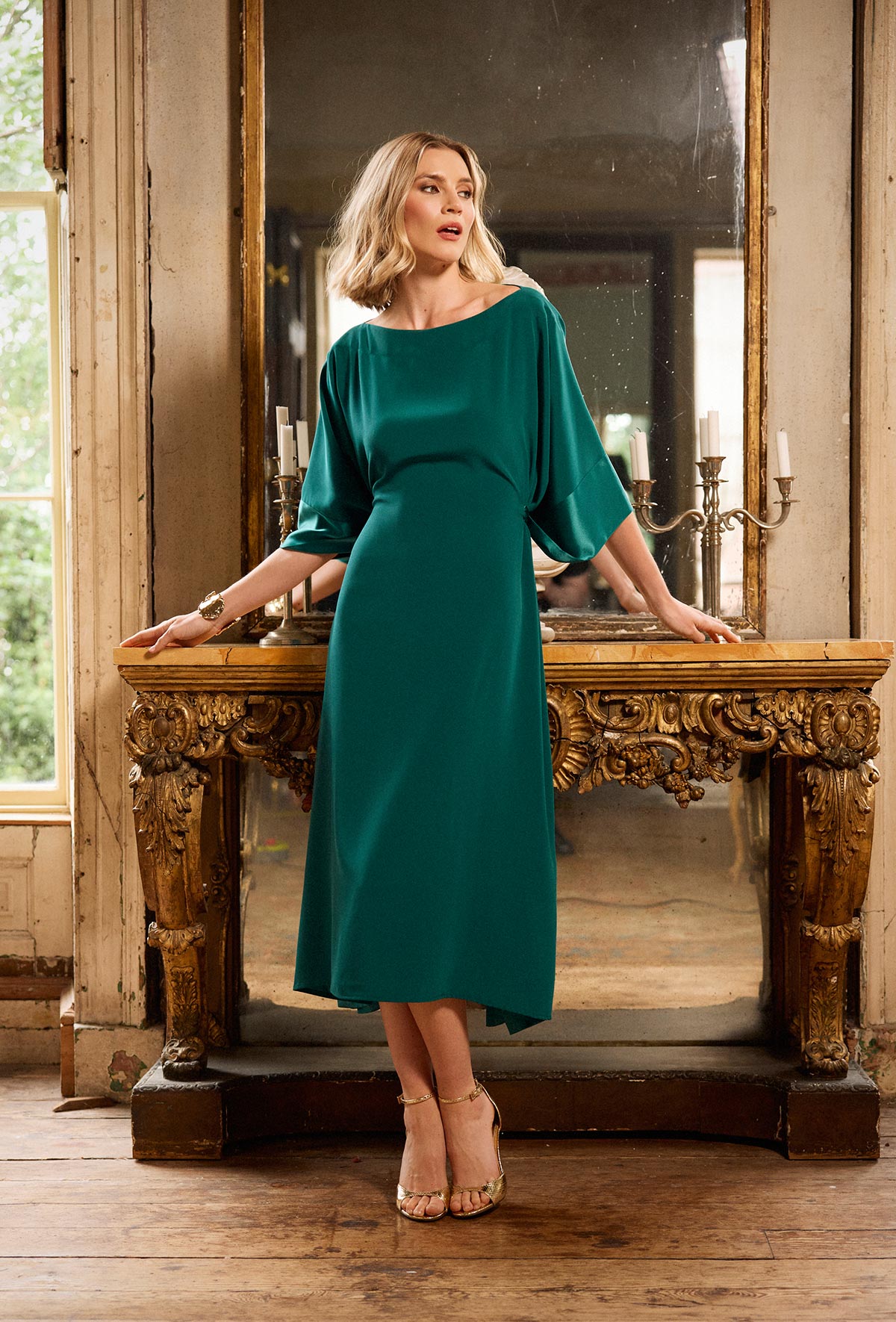 Woman in a teal dress standing in a room with wooden furniture and large mirror.