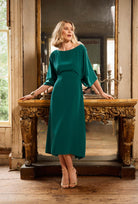 Woman in a teal dress standing in a room with wooden furniture and large mirror.