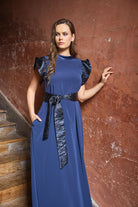 A woman wearing a long royal blue dress with dark metallic cap sleeves and a matching dark belt, standing against a textured reddish-brown wall next to wooden stairs.