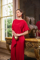 A woman wearing a vibrant red draped dress and holding a gold clutch stands in an ornate room with classical busts and a large window.