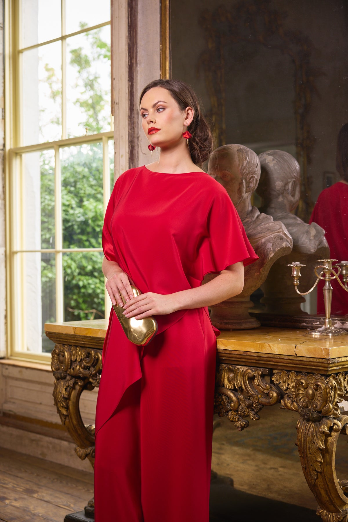 A woman wearing a vibrant red draped dress and holding a gold clutch stands in an ornate room with classical busts and a large window.