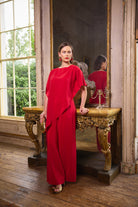 A woman models a vibrant red draped jumpsuit in a vintage room with wooden floors, an ornate gold console table, and a large mirror reflecting her back.