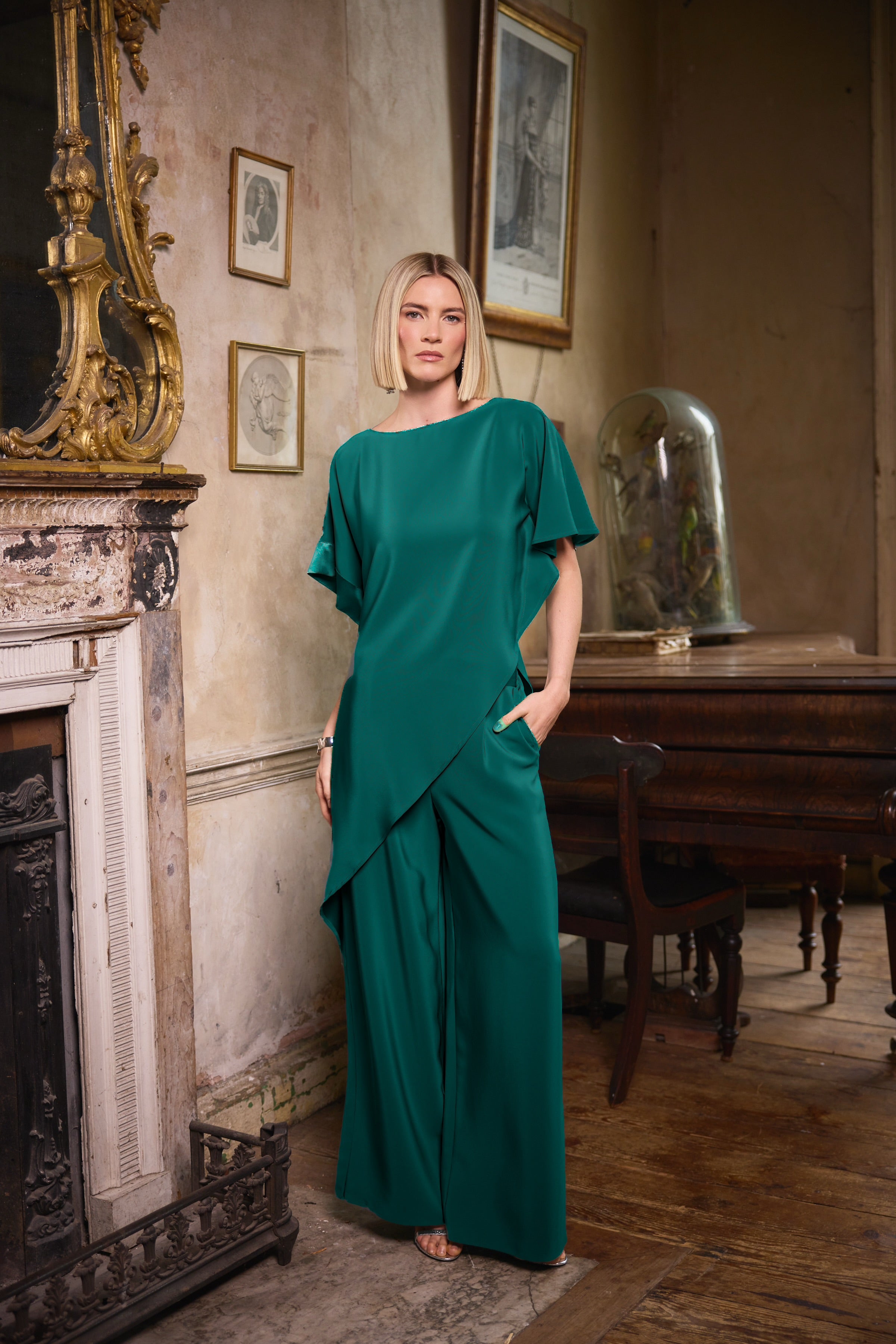 A woman stands in a vintage room wearing a flowing emerald green draped jumpsuit with wide-leg pants and short flutter sleeves. The room features a fireplace, framed pictures, and a wooden table in the background.