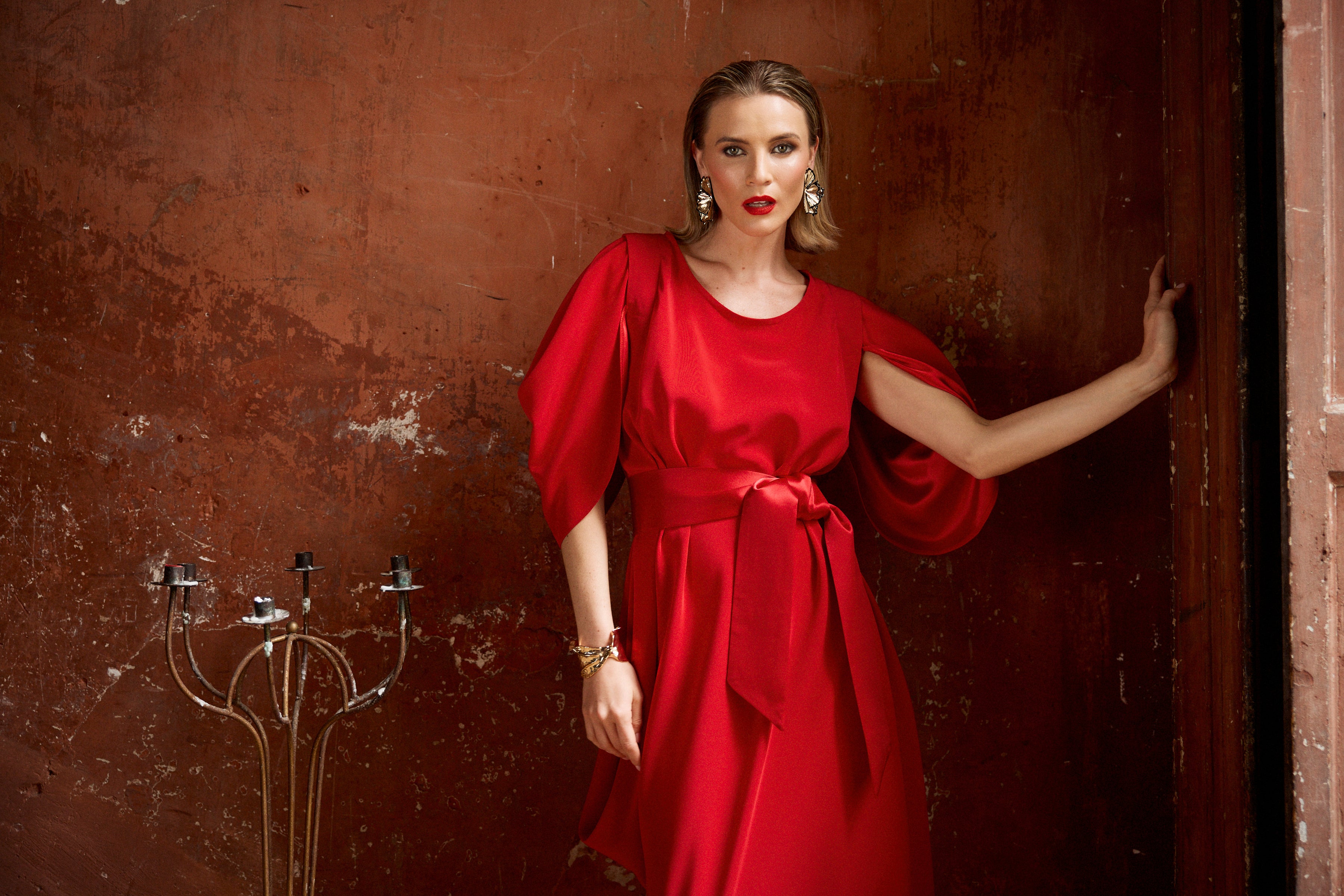 Woman in a red dress standing against a textured brown wall.