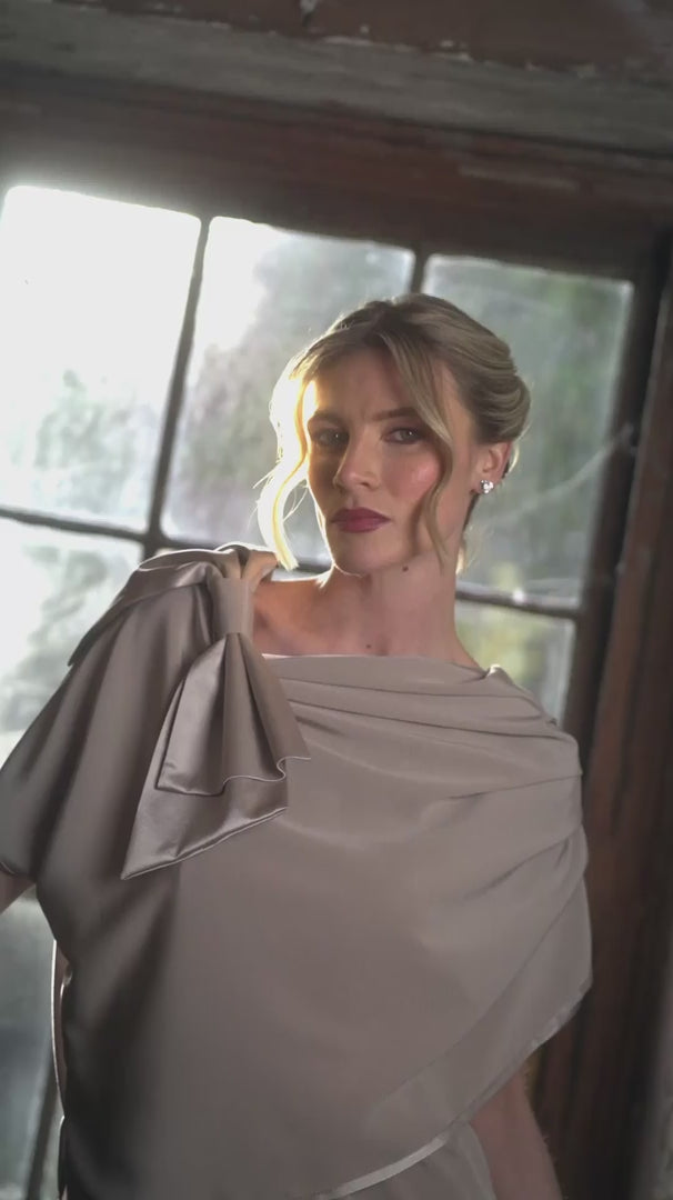 Woman in a beige dress standing in a rustic room with wooden floors and a large window.